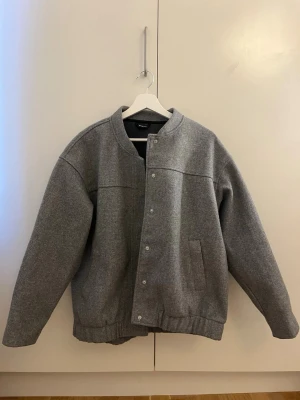 Grå bomberjacka från Gina Tricot - Snygg grå bomberjacka från Gina Tricot med minimalistisk design. Jackan har tryckknappar framtill, två sidofickor och ribbad nederkant. Tillverkad i ett mjukt, filtliknande material som ger en clean och modern look.