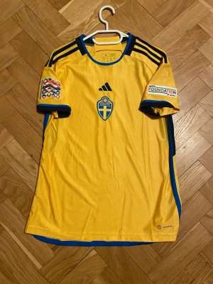 Svensk landslagströja Adidas gul - Gul matchtröja från Adidas med Sveriges landslagsemblem på bröstet och svarta detaljer på axlarna. Blåa inslag vid ärmslut och sidor, Nations League-märke på ena ärmen och Foundation-märke på andra. Tillverkad i lätt funktionsmaterial, perfekt för fotboll.