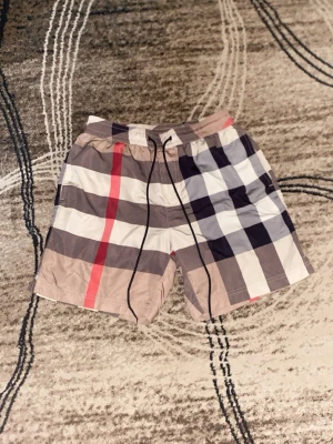 Säljer burbberry short för sommaren - Säljer ett par klassiska short för sommaren, från Burberry med stort rutmönster i beige, svart, vitt och rött. Shortsen har snörning i midjan och är tillverkade i ett lätt syntetmaterial som torkar snabbt. Perfekta för sommaren!