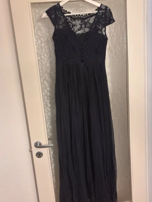 Marinblå långklänning med spets - Elegant marinblå långklänning med v-ringning och spetsdetaljer upptill. Klänningen har genombruten spets vid axlar och rygg, samt knappar framtill på överdelen. Kjoldelen är i ett tunt, flytande material som ger ett mjukt fall.