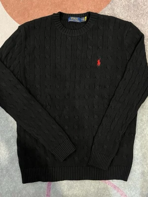 Svart kabelstickad tröja Polo Ralph Lauren - Svart kabelstickad tröja från Polo Ralph Lauren med rund hals och lång ärm. Tröjan har ett klassiskt rött broderat logotyp på bröstet och ribbade muddar vid ärmslut och nederkant. Perfekt för dig som gillar stilrena och tidlösa plagg.
