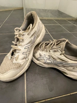 Vita Asics sneakers med meshdetaljer - Snygga vita sneakers från Asics med klassisk logga på sidorna. Skorna har meshpaneler för extra andningsförmåga och detaljer i syntetmaterial. Perfekta för dig som gillar sportig stil och vill ha bekväma skor till vardags.