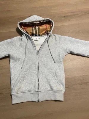 Grå zip hoodie från Burberry - Snygg grå zip hoodie från Burberry med klassiskt rutigt foder i huvan. Hoodien har dragsko i huvan, två fickor fram och ribbade muddar. Perfekt för dig som vill ha en stilren och exklusiv look med en twist av Burberrys ikoniska mönster.