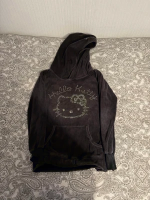 Mjuk Hello Kitty hoodie från H&M - Säljer en svart velour-hoodie från H&M med Hello Kitty-motiv i glittriga stenar på bröstet. Hoodien har stor huva och magficka, samt långa ärmar med muddar. Perfekt för dig som gillar gulliga detaljer och vill ha något riktigt bekvämt.