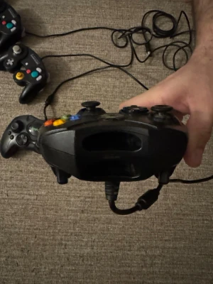 Svart Xbox handkontroll med sladd - Klassisk svart Xbox handkontroll med sladd, ergonomisk form och tydliga färgade knappar. Tillverkad i plast med två analoga spakar, styrkors och triggers. Perfekt för retrospel på original Xbox.