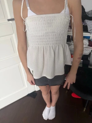 Vit smockad linnetopp med knytband - Supersöt vit linnetopp med smockad överdel och tunna axelband med knyt. Toppen har en lätt volang nedtill och är perfekt till sommaren. Luftigt material och enkel att matcha med jeans eller kjol.