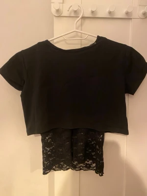 Svart croppad topp med spetsdetaljer - Tvådelad svart topp med en croppad t-shirt i bomull och en ärmlös spets-topp under. Spetsen har blommigt mönster och ger en snygg lager-på-lager-look. Perfekt för dig som gillar att sticka ut med detaljer och kontraster.