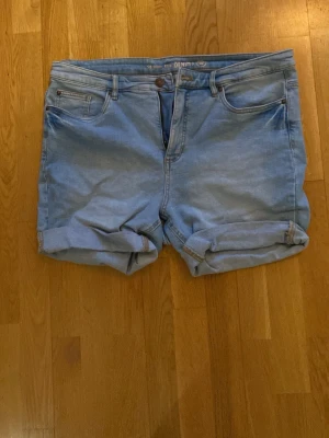 Ljusblå denimshorts från Ella - Säljer ett par ljusblå denimshorts från Ella med klassisk femficksdesign och uppvikta benslut. Shortsen har normal passform och stängs med knapp och dragkedja. Perfekta till sommaren för en avslappnad look.