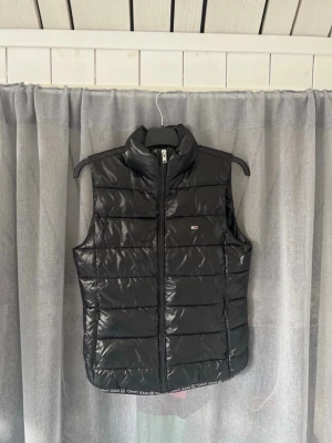 Svart väst från Tommy Jeans - Snygg svart väst från Tommy Jeans med quiltad design och hög krage. Västen har dragkedja framtill och logotyp på bröstet samt textdetalj längs nederkanten. Perfekt för lager-på-lager och ger en cool look.