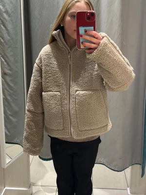 Beige teddyjacka med stora fickor - Supermjuk och fluffig beige teddyjacka med dragkedja framtill och två stora fyrkantiga fickor. Jackan har hög krage! Perfekt till våren/hösten.