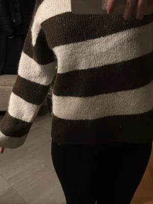 Brun och vit randig stickad tröja - Mysig stickad tröja med breda bruna och vita horisontella ränder. Tröjan har lång ärm och en avslappnad passform. Perfekt för dig som gillar en enkel men trendig look. Materialet ser fluffigt och mjukt ut, vilket gör den extra skön att bära. 