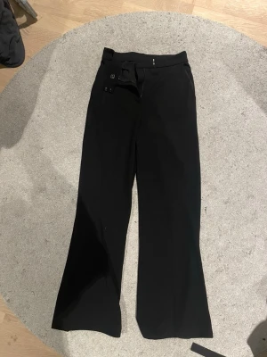 Svarta vida kostymbyxor - Säljer ett par svarta kostymbyxor med hög midja och vida ben. Byxorna har diskreta sidofickor och stängs med hake och knapp i midjan. Klassisk och stilren look som passar till många olika outfits.