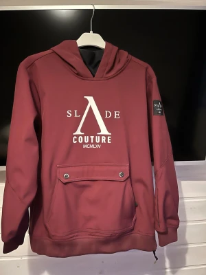 Slade jacka - Vinröd skidjacka från slade med stor ficka på magen. Ficka på ena armen för skidpass, passform som en hoodie men finns drajskedja för att få på sig enklare.