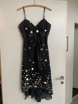 Svart paljettklänning med rosett från Forever Yours - Festklänning från Forever Yours International Corp., ett brud- och festklädesföretag som tidigare var baserat i New York. Svart sidenliknande klänning med smala axelband, v-ringning och stor rosett i midjan. Klänningen har asymmetrisk nederkant och är dekorerad med stora, glittriga paljetter i silver. Perfekt för dig som vill sticka ut på festen.