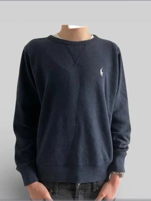 Mörkblå sweatshirt från Polo Ralph Lauren - Klassisk mörkblå sweatshirt från Polo Ralph Lauren med rund halsringning och broderad vit logga på bröstet. Tröjan har ribbade muddar vid ärmslut och nederkant samt en snygg detalj vid halsen. Perfekt för en avslappnad och stilren look. Har några fläckar men syns inte jättetydligt, kanske kan gå att få bort några
