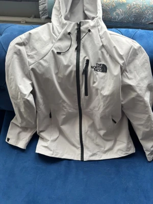 Grå vindjacka The North Face L - Snygg grå vindjacka från The North Face Summit Series med huva, svarta detaljer och logga på bröstet. Jackan har dragkedja framtill, bröstficka med dragkedja och justerbara ärmslut. Perfekt för att hålla vinden borta under äventyr.