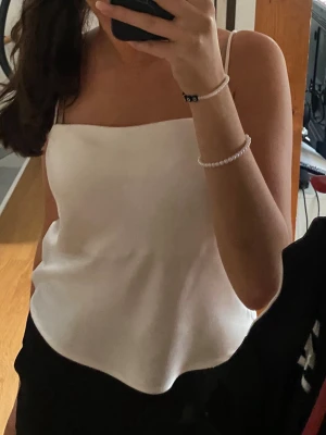 Vit satin linnetopp med smala band - Snygg vit linnetopp i satin med smala axelband och rak halsringning. Perfekt att styla med jeans eller kjol för en trendig look. Tunn och lätt, ger en elegant känsla och passar till många olika outfits.