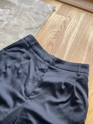 Svarta vida kostymbyxor i satin - Svarta kostymbyxor med hög midja och vida ben i ett glansigt material. Små ”revor” i materialet (se bilderna). 