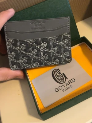 Grå plånbok kortfodral från Goy4ard - Snyggt grått kortfodral från Goy4ard med klassiskt mönster i vitt och svart. Fodralet har flera kortfack och är tillverkat i canvas och läder. Perfekt för dig som vill ha en stilren och exklusiv accessoar i fickan.