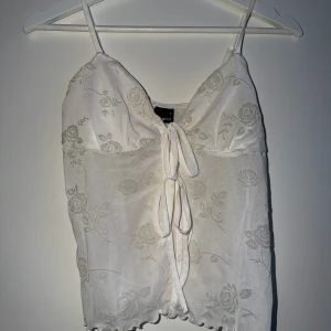 Vit blommig topp med knyt - Söt vit topp från Gina tricot med tunna axelband och blommor. Toppen har dekorativ knytning framtill och lätt volangkant nedtill. Perfekt för sommarens alla tillfällen och ger en romantisk vibe.