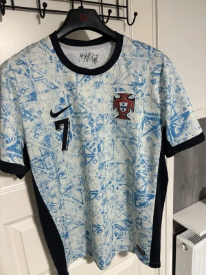 Portugal Ronaldo Nike fotbolls Jersey - Säljer en Portugal fotbollströja från Nike i ljusblått och vitt med abstrakt mönster. Tröjan har svarta detaljer, nummer 7 på bröstet och Portugals landslagsemblem. Perfekt för dig som älskar fotboll och vill sticka ut på planen.