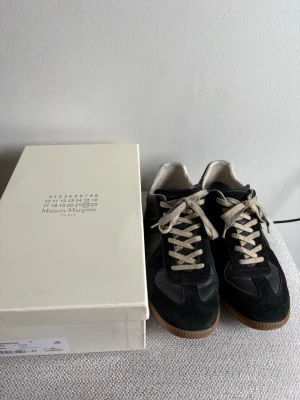 Maison Margiela svarta sneakers - Maison Margiela sneakers i svart läder och mocka med beige snörning och gummisula. Klassisk låg modell med stilren design och diskreta detaljer. Perfekt för dig som gillar exklusiva och trendiga skor.