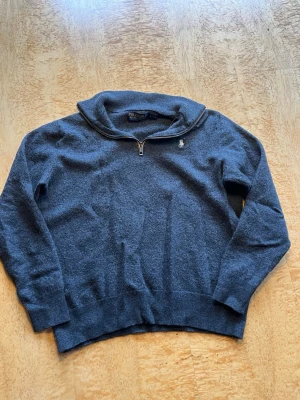 Grå halvzip tröja från Polo Golf - Grå stickad tröja från Polo Golf Ralph Lauren med halvzip och liten broderad logga på bröstet. Tröjan har ribbade muddar och krage, perfekt för lager på lager. Materialet känns mjukt och håller värmen bra. Därimot har tröjan krympt i tvätten så storleken är inte helt rätt men den borde passa på någon runt 150-160