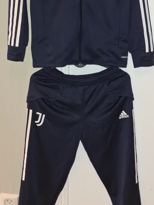 Juventus träningsset från Adidas - Mörkblått träningsset från Adidas med Juventus-logga. Setet består av en zip-jacka och matchande byxor, båda med vita ränder längs ärmar och ben. Tillverkat i ett lätt och stretchigt material som passar perfekt till fotbollsträning eller gym. St 152 passar barn 11-12 åring