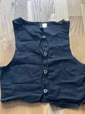 Svart väst med hjärtknappar - Svart väst från Divided med fyra dekorativa hjärtformade knappar framtill. Modellen är croppad.