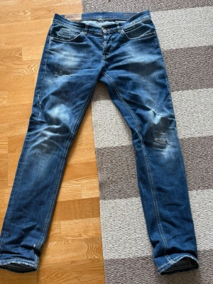 Dondup jeans  - Säljer ett par blå jeans med slitningar och tvättade detaljer. Jeansen har fem fickor, raka ben och kontrastsömmar. Snyggt slitna partier på knän och lår samt lite färgstänk för en cool, avslappnad look. Perfekta för dig som gillar streetstyle. Skriv vid funderingar eller frågor✌🏼