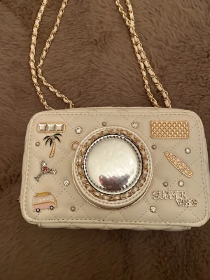 Krämvit handväska med spegel och kedja från Aldo  - Unik krämvit handväska i quiltat material med guldfärgad kedjerem. Väskan har dekorativa detaljer som palmer, surfbräda, van och texten 'SUMMER VIBES'. Framsidan har en stor spegel med guldram som kan öppnas. Perfekt för dig som gillar statement-accessoarer. Från Aldo