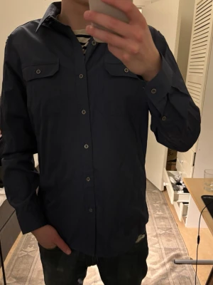 Mörkblå skjorta med bröstfickor - Snygg mörkblå skjorta med två bröstfickor och knappar hela vägen framtill. Klassisk krage och långärmad modell, perfekt för en clean och stilren look. Tillverkad i bomull för skön känsla.