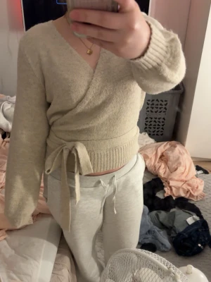 Beige omlottstickad tröja - Mysig beige stickad omlottopp med knytband i midjan och bred ribbad kant. Tröjan har långa ärmar och en djup v-ringning. Perfekt för lager på lager och enkel att styla till olika outfits.