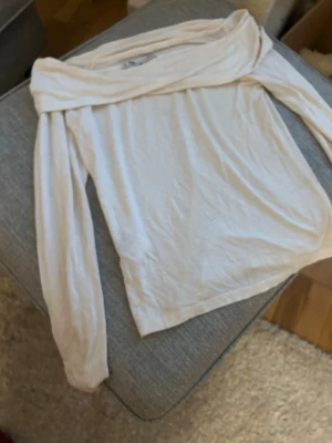 Vit offshoulder topp med långa ärmar - Säljer en stilren vit offshoulder topp med långa ärmar. Toppen har en bred, vikt kant över axlarna som ger en snygg och trendig look. Perfekt att matcha med jeans eller kjol för en cool outfit.