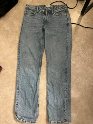 Blå raka jeans med rå kant - Säljer ett par klassiska blå jeans med rak passform och rå kant nedtill. Jeansen har fem fickor, knappgylf och är tillverkade i denim. Perfekta för en avslappnad och trendig look.