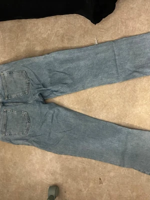 Ljusblå raka jeans med slitning - Säljer ett par ljusblå jeans med raka ben och stor slitning på ena låret. Jeansen har klassiska bakfickor och är tillverkade i denim. Perfekta för en avslappnad och trendig look.