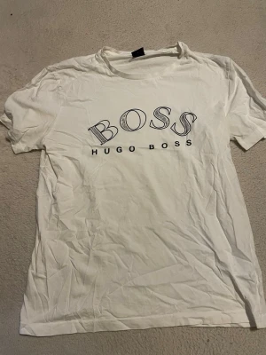 Vit Hugo Boss t-shirt med logga - Vit t-shirt från Hugo Boss med stor BOSS-logga och texten HUGO BOSS på bröstet. Klassisk passform och rund halsringning. Perfekt för en clean och stilren look.