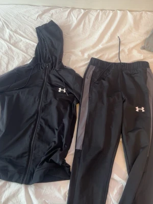 Svart träningsset från Under Armour båda är strl S - Snyggt svart träningsset från Under Armour med zip-hoodie och matchande byxor. Setet har gråa detaljer längs sidorna och vita loggor på både jacka och byxor. Tillverkat i ett mjukt och stretchigt funktionsmaterial som passar perfekt till träning.