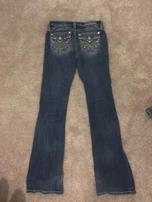 Mörkblå bootcut jeans med broderi - Säljer ett par mörkblå bootcut jeans med snygga broderade bakfickor och dekorativa nitar. Jeansen har en klassisk femficksmodell och detaljer i silver på fickorna. Perfekta för dig som gillar unika detaljer och en avslappnad stil.