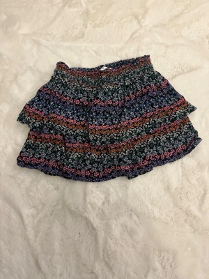 Blommig kortkjol från M&S - Söt kortkjol från M&S med blommigt mönster i flera färger som lila, orange, blå och vit. Kjolen har volangdetaljer och resår i midjan för en bekväm passform. Perfekt för dig som gillar färgglada och lekfulla plagg.