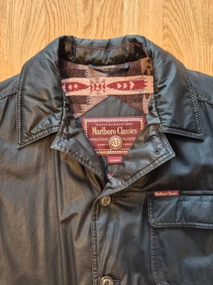 Vintage Marlboro Classics herrjacka, stl XL - Vintage Marlboro Classics jacka med ett unikt mönstrat foder. Modellen är från mitten till slutet på 1990-talet. Jackan har en klassisk krage och knäppning framtill. Den är utrustad med flera praktiska fickor, inklusive två bröstfickor med lock och två större fickor nedtill, också med lock. En innerficka finns för förvaring av värdesaker. Designen kombinerar funktionalitet med en distinkt stil. 