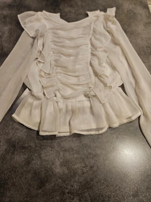 Lindez  - Snygg vit blus i tunt, transparent tyg med volangdetaljer framtill och längs sidorna. Intw använt den så mycket. Stolek 146 ifrån Lindez.