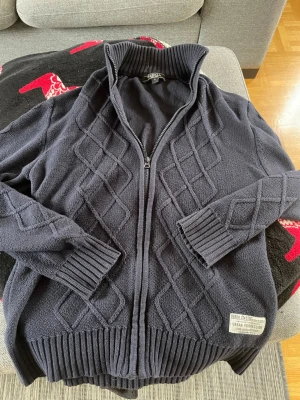 Mörkblå stickad ziptröja från Berga Urban Profession - Mörkblå stickad ziptröja från Urban Profession med hög krage och ribbade muddar. Tröjan har ett snyggt diamantmönster framtill och en patch vid nederkanten. Perfekt för lager på lager och chill stil. Priset kan diskuteras. 