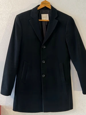 Svart klassisk ullkappa - Stilren svart ullkappa från Total Studio med tre knappar framtill, klassisk krage och två snedställda fickor. Passar  perfekt till höstens och vinterns outfits.