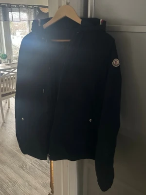Svart vindjacka från Moncler - Snygg svart vindjacka från Moncler med huva och dragkedja framtill. Jackan har två fickor med tryckknappar och Moncler-logga på ärmen. Perfekt för blåsiga dagar och enkel att matcha med olika outfits. Obs rep och NFC scan funkar exakt som den ska. 