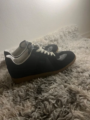 Maison margela - Snygga svarta sneakers med vita skosnören och kontrasterande brun gummisula. Ovandelen är i en mix av mocka och slätt läder, med klassisk låg profil och vit insida. Perfekta för en clean och stilren look.