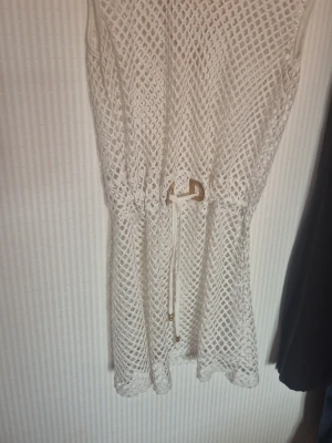 Vit strandklänning med nätmönster - Säljer en vit strandklänning i nätmönstrad mesh med knytband i midjan och guldfärgade detaljer. Perfekt att dra över bikinin på stranden. Klänningen är ärmlös och har en avslappnad passform som ger en luftig känsla.