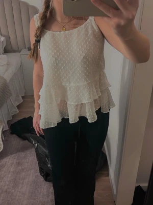 Vit volangtop med prickigt mönster - Söt vit topp med volanger och prickigt mönster. Toppen är ärmlös och har dubbla lager volanger nedtill för en flowy look. Materialet känns lätt och luftigt, perfekt för varmare dagar. Passar till både jeans och kjol för en trendig stil.