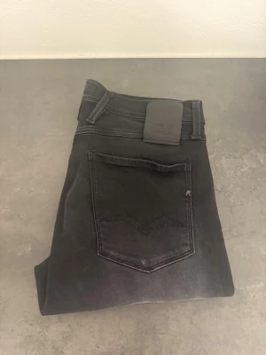Svarta Replay Anbass jeans stl 31/32 - Snygga svarta slim fit jeans från Replay med klassisk femficksmodell och diskret Replay-logga på bakfickan. Jeansen har en modern passform och är tillverkade i stretchigt denimtyg för extra komfort. Perfekta för dig som gillar en stilren och enkel look.
