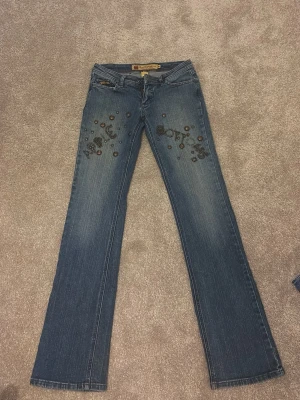 Blå jeans med nitar från Apple Bottoms - Säljer ett par blå bootcut jeans från Apple Bottoms med coola nitar och dekorationer på framsidan. Jeansen har klassisk femficksmodell, dragkedja och knapp, samt detaljerad etikett i midjan. Materialet är 98% bomull och 2% spandex för skön stretch. OPS- dom saknar en knapp, det hade kostat 100kr att fixa det så jag säljer dom för 450kr istället för 550kr